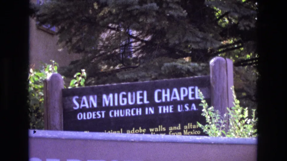 1973 New Mexico home-movie frame showing a sign for San Miguel Chapel beside trees, shrubs, and the edge of the chapel grounds.