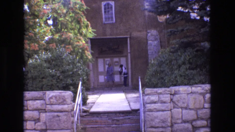 1973 New Mexico home-movie frame showing two adult women at the San Miguel Chapel doorway, framed by adobe walls, greenery, stonework, and the front steps.