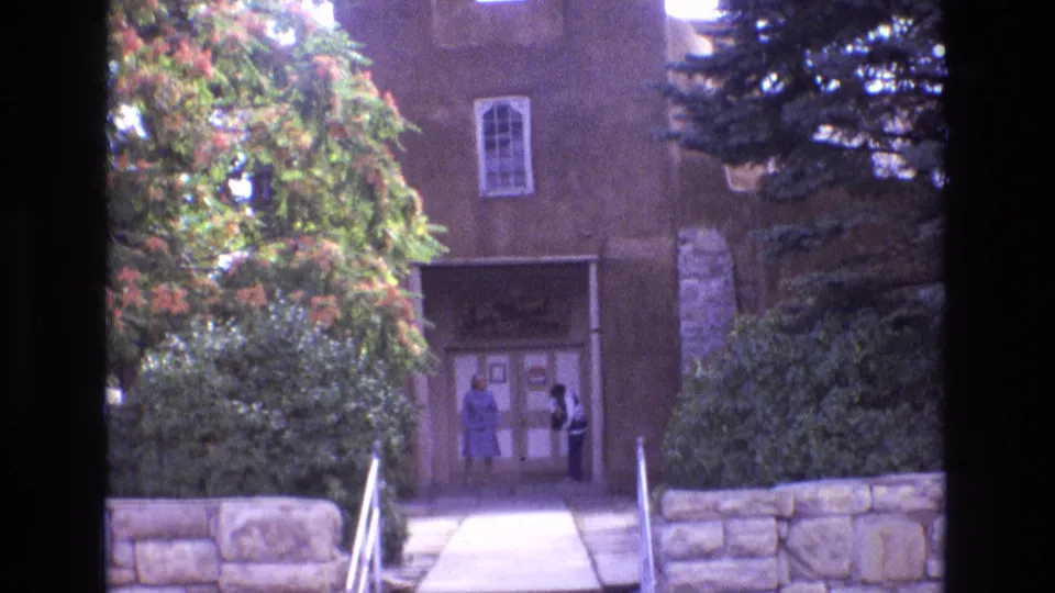 1973 New Mexico frame of San Miguel Chapel with two adult women at the entrance, a small window above, stone wall below, and trees partly obscuring the adobe facade.