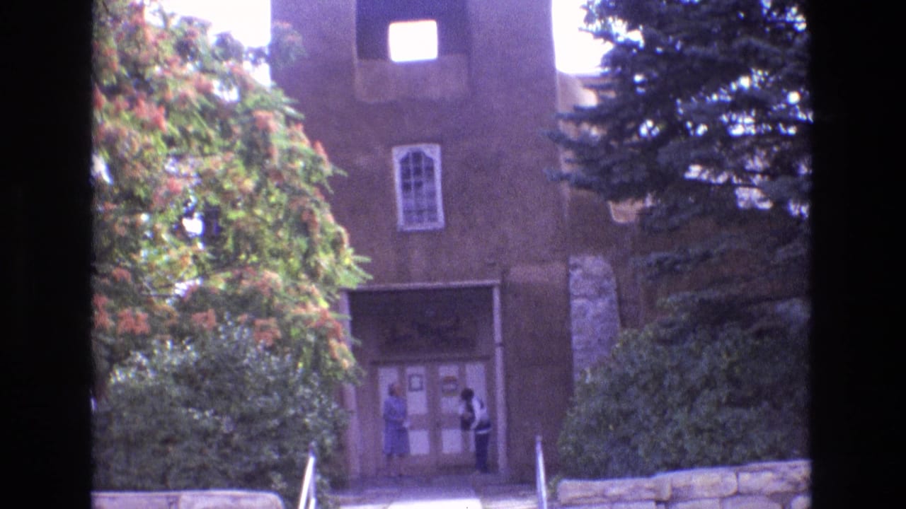 1973 New Mexico 8mm frame showing a historic adobe church exterior with a bell tower opening, arched window, trees, and two visitors by the entrance.