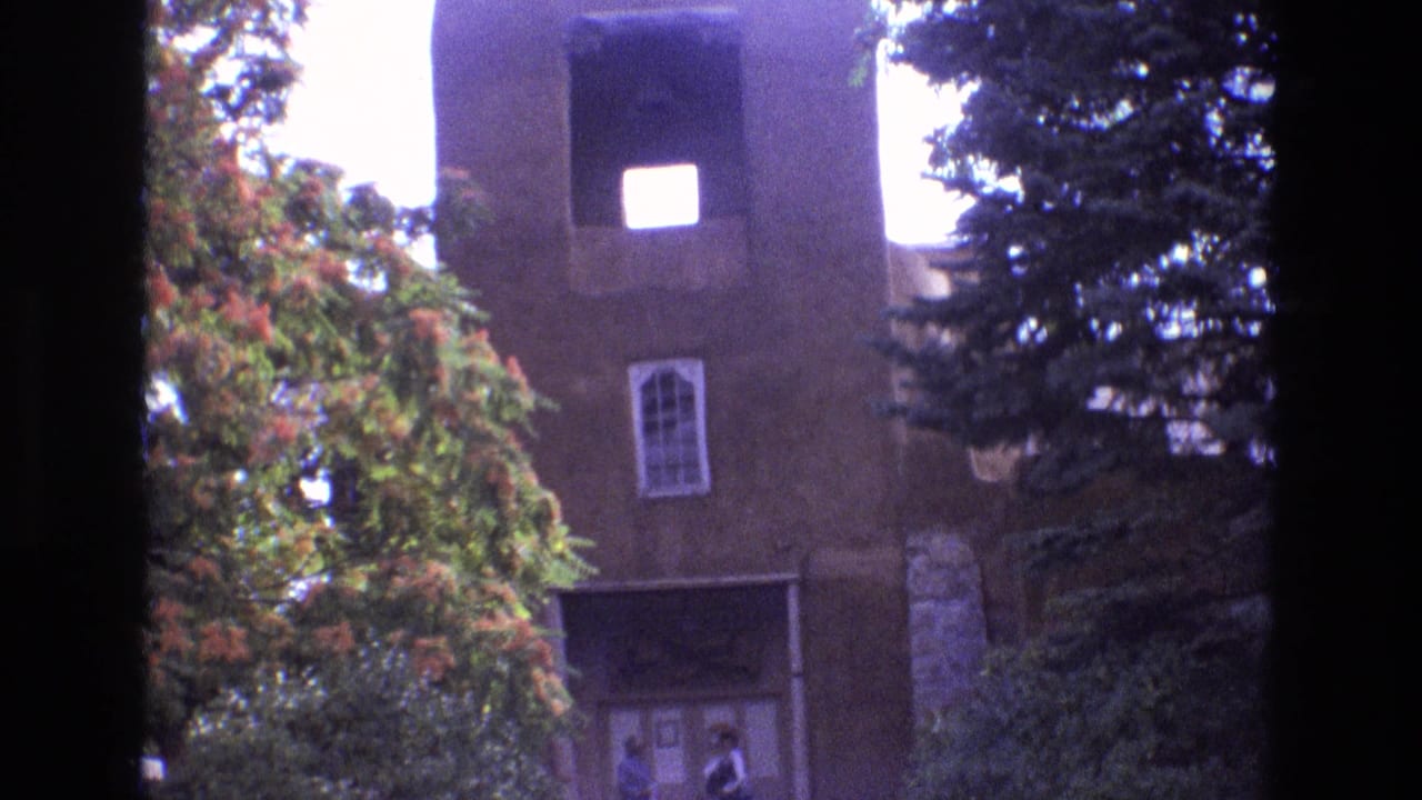 1973 New Mexico home-movie frame of an adobe church facade, bell tower, arched window, and visitors near the shaded entrance, framed by dense trees.