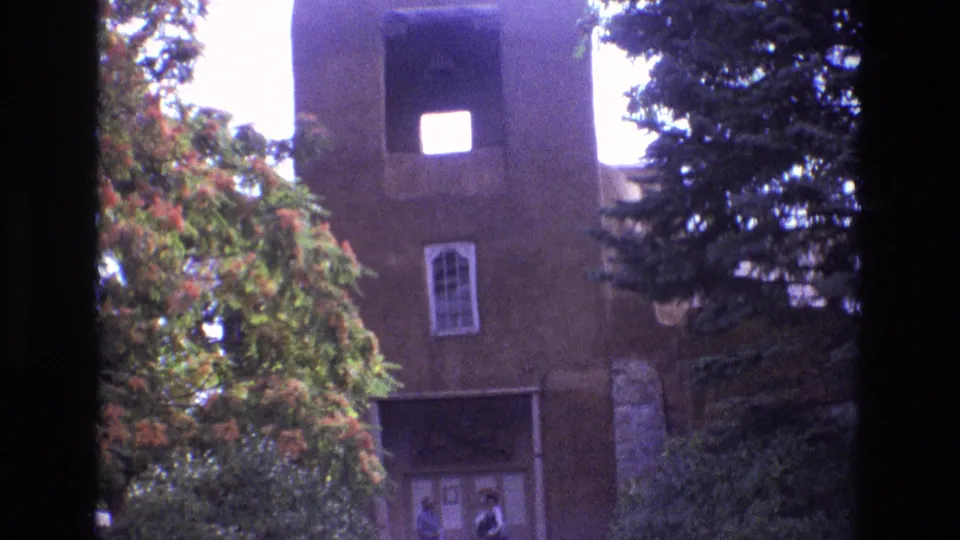 1973 New Mexico home-movie frame of an adobe church facade, bell tower, arched window, and visitors near the shaded entrance, framed by dense trees.