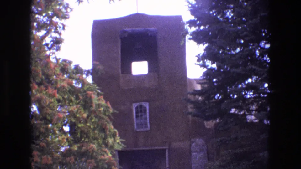 1973 New Mexico 8mm frame with the adobe church tower, bright bell opening, arched window, leafy trees, and part of the entrance below.