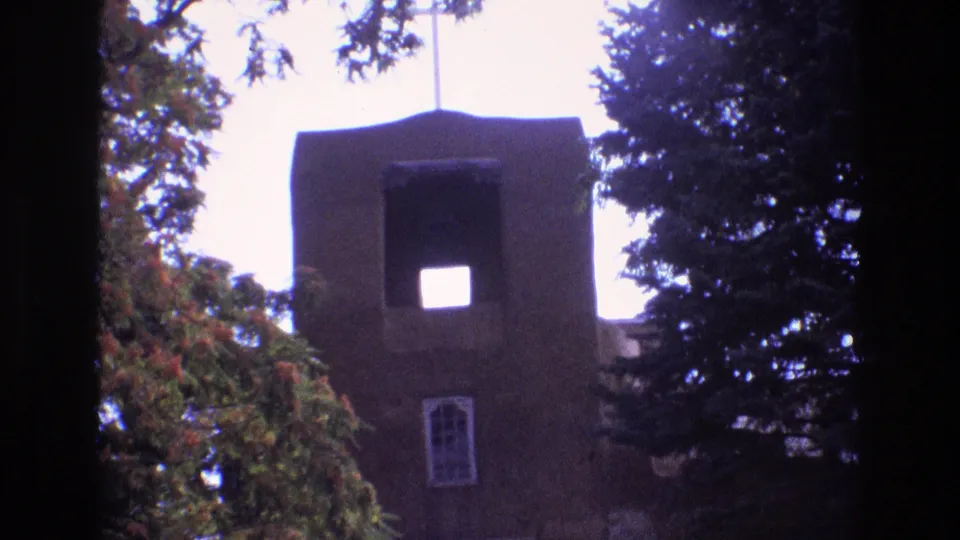 1973 New Mexico home-movie frame focusing on the adobe church bell tower, arched window, stucco wall, surrounding trees, and high-contrast sky.