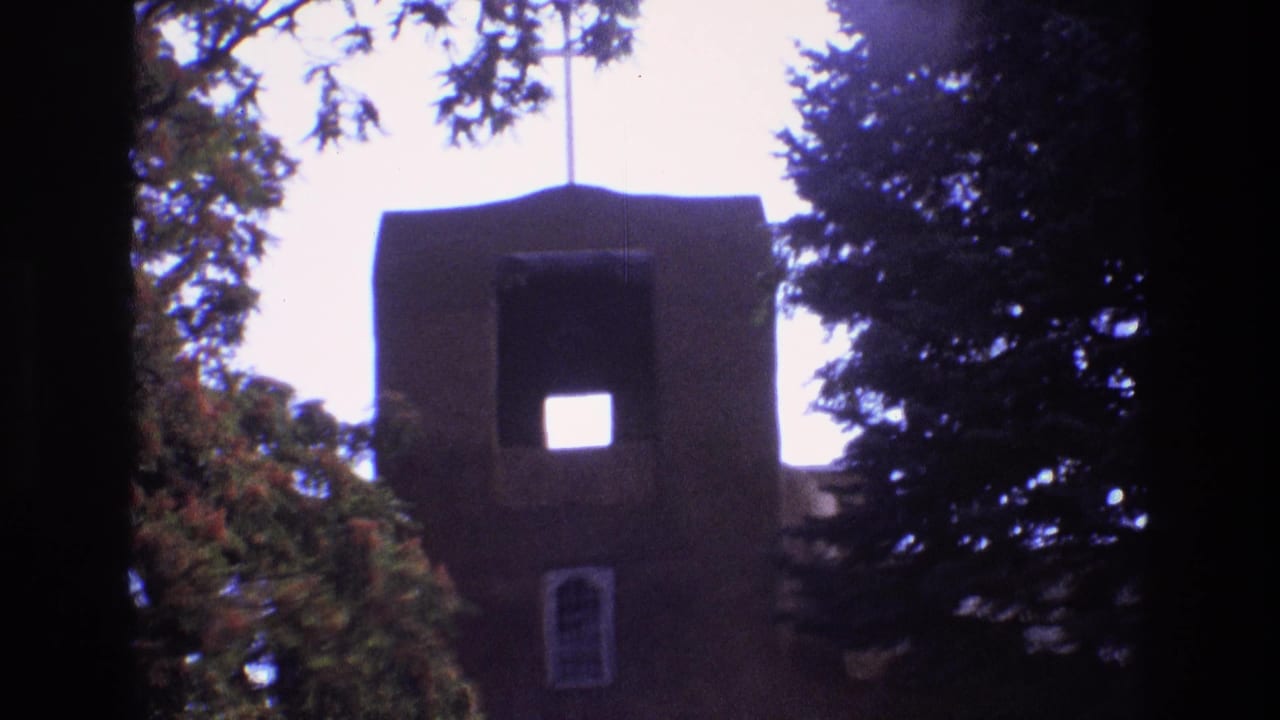1973 New Mexico 8mm frame showing a brown adobe-style church tower with a square upper opening, a small arched window, and trees partly blocking the view.