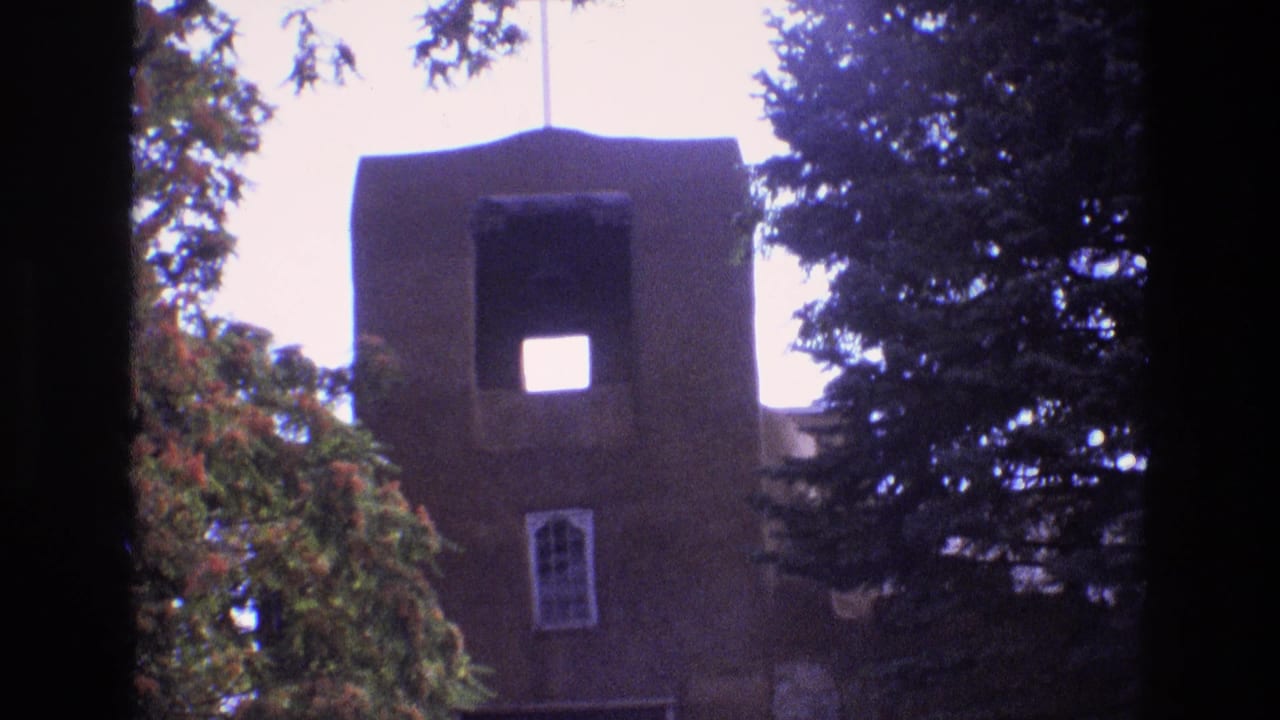 1973 New Mexico 8mm frame of a tall brown adobe-style church tower with a cross above it, a square opening near the top, an arched window below, and trees on both sides.
