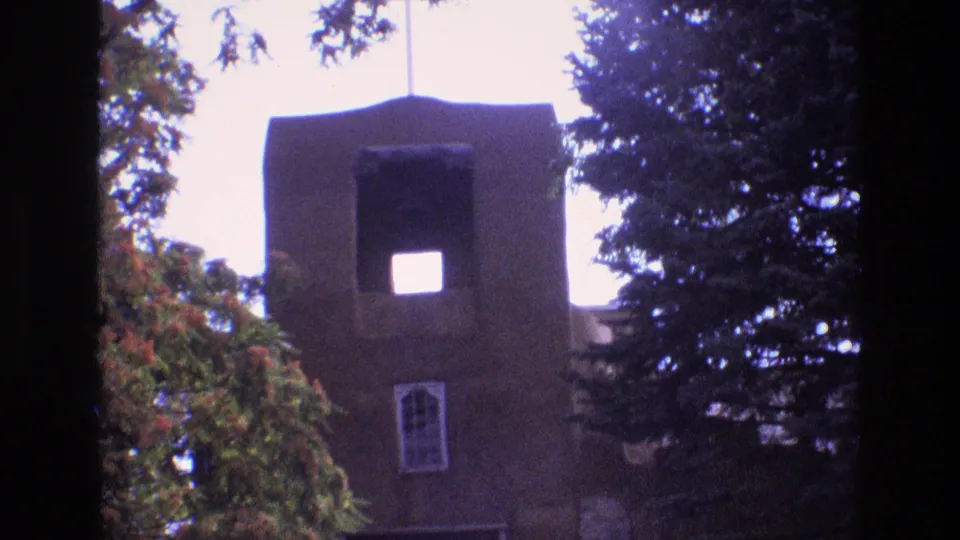 1973 New Mexico 8mm frame of a tall brown adobe-style church tower with a cross above it, a square opening near the top, an arched window below, and trees on both sides.