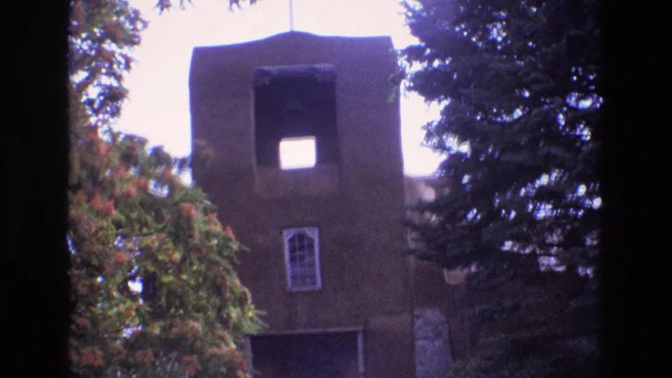 1973 New Mexico 8mm frame showing the brown church tower from slightly farther back, with a square upper opening, arched window, and leafy trees filling the foreground edges.