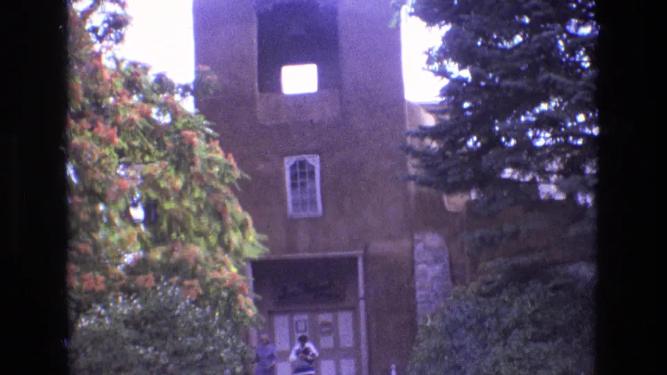 1973 New Mexico 8mm frame revealing more of the brown adobe-style church facade, including the tower, cross, arched window, doorway, shrubs, and two people standing near the entrance.