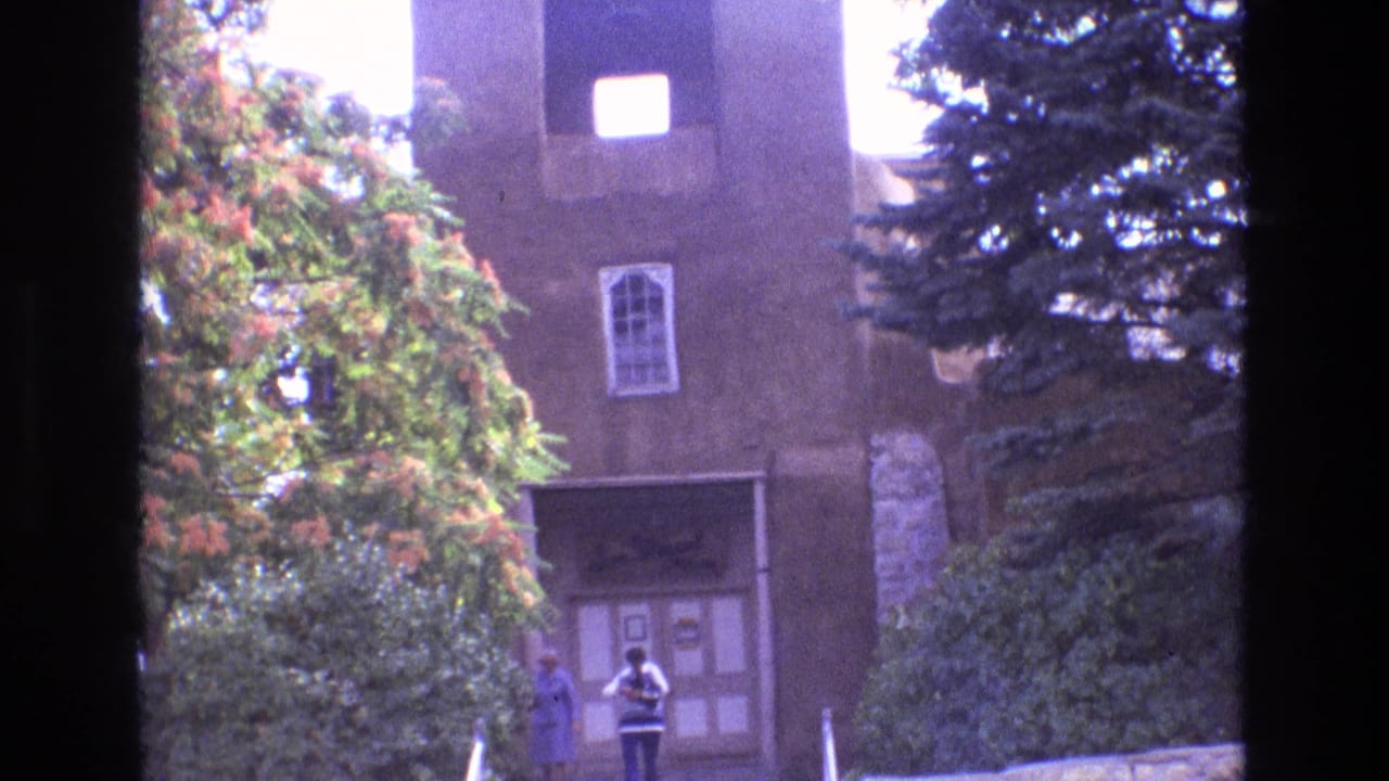 1973 New Mexico home-movie frame showing a restored-looking adobe mission entrance with bell openings, stone steps, trees, and two visitors near the doorway.