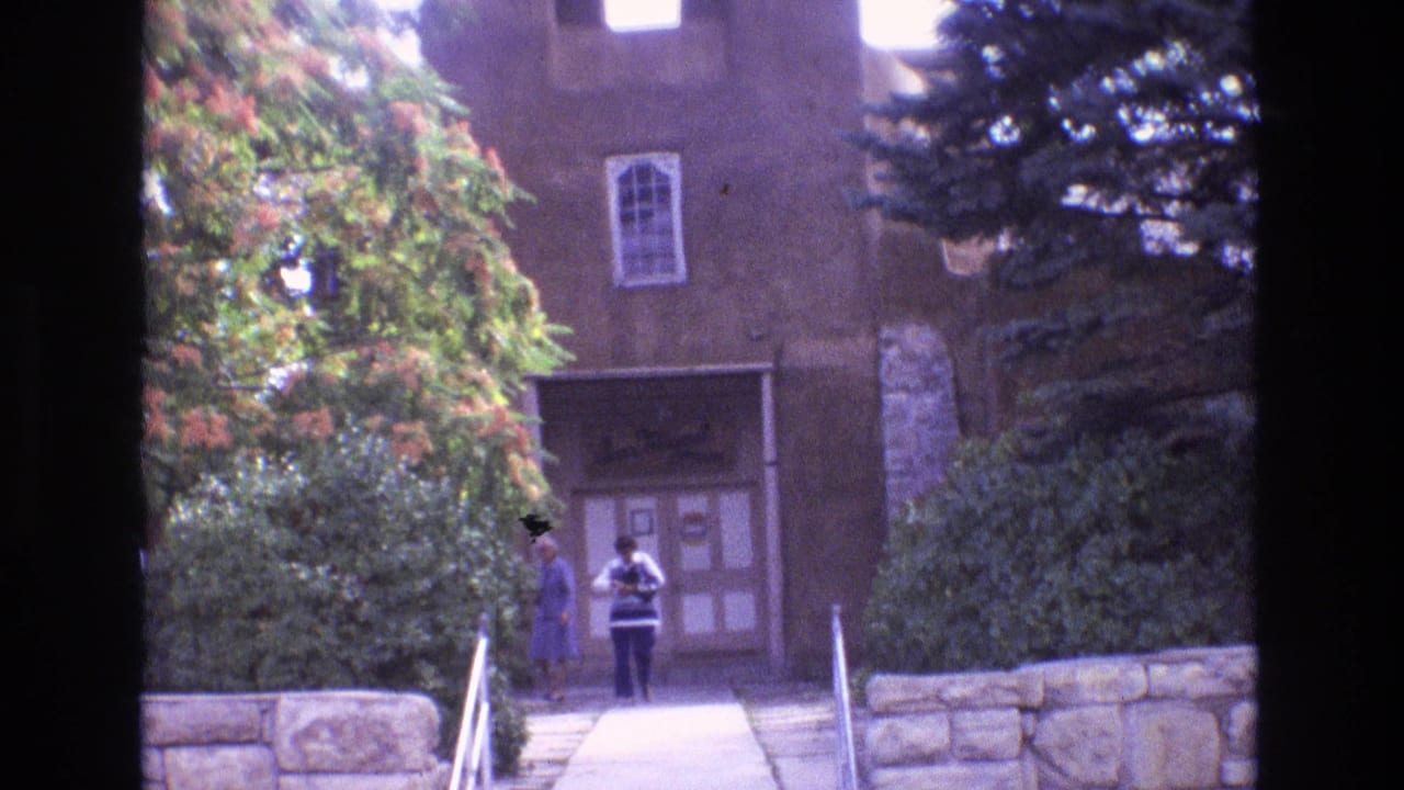 Adobe church entrance with trees, stone wall, walkway, and two people near the doorway.