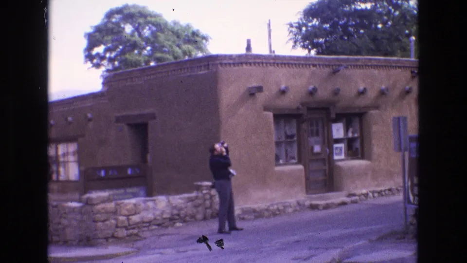 1973 New Mexico home-movie frame showing a man photographing a low adobe building with a flat roof, wooden doors, windows, stone wall, and nearby trees.