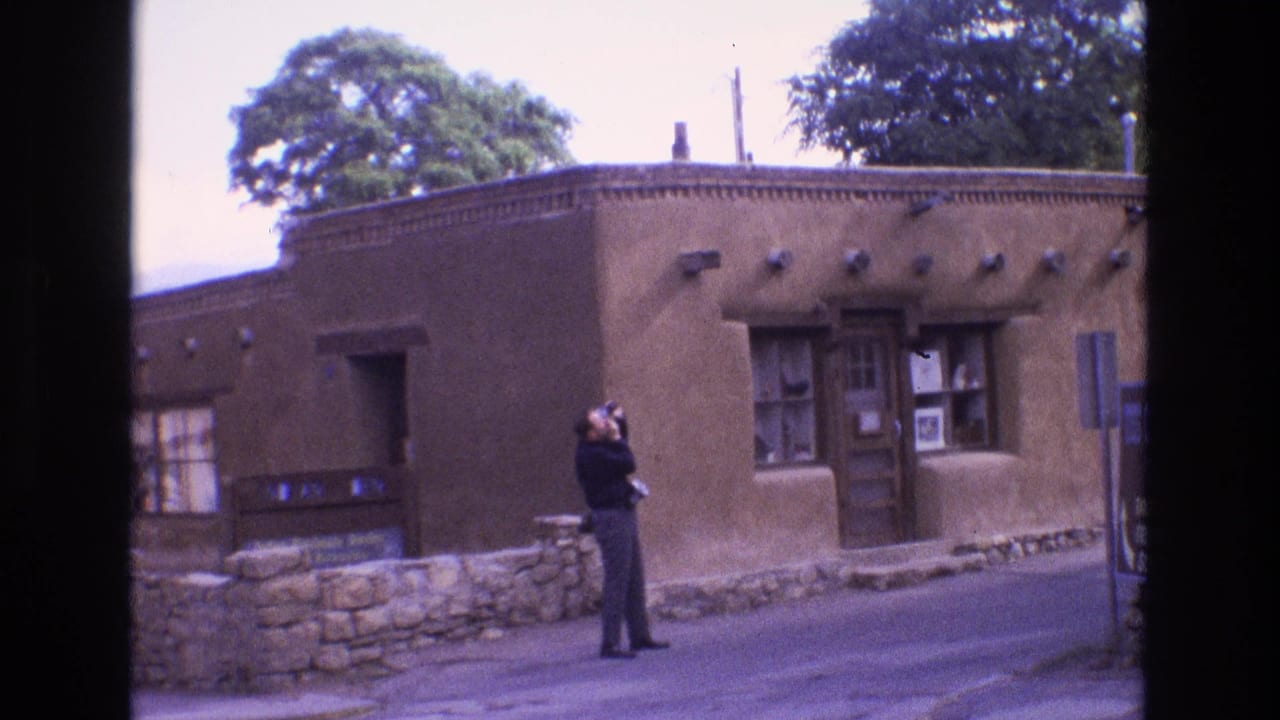 In 1973 New Mexico home-movie footage, a man in dark clothing raises a camera while standing before a brown adobe-style building with a stone wall and wooden door.