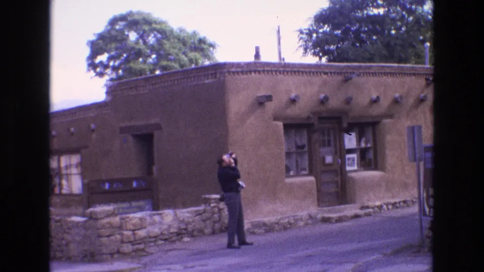 In 1973 New Mexico home-movie footage, a man in dark clothing raises a camera while standing before a brown adobe-style building with a stone wall and wooden door.