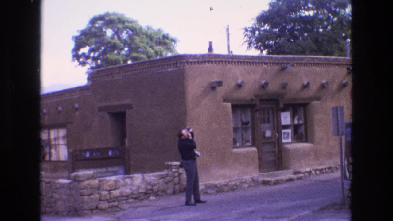Man aims a camera upward near a brown adobe-style building and stone wall.