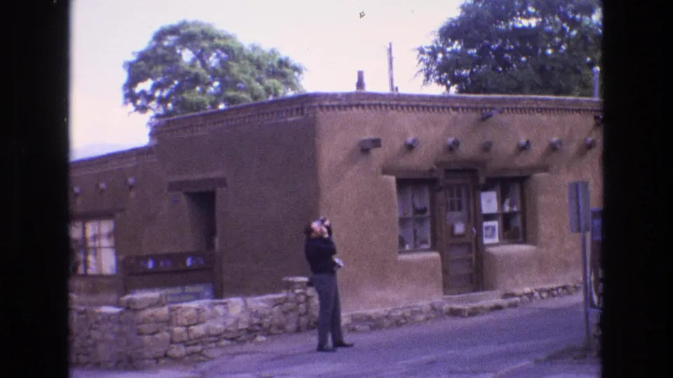 The man continues aiming his camera upward from the street in front of a New Mexico adobe-style exterior, with windows, door, stonework, and trees visible.
