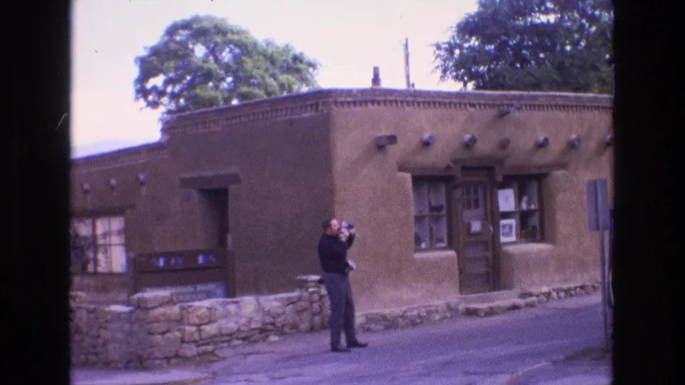 A 1970s home-movie frame shows the man near a low stone wall, holding a camera toward the adobe-style facade with a wooden door and shaded windows.