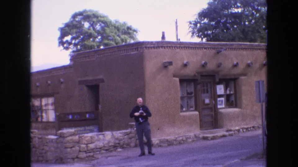 At the end of the clip, the man faces more toward the camera with his camera lowered in front of the brown adobe-style building and stone wall.