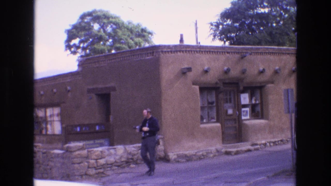 1973 New Mexico home-movie frame showing an adult man in dark clothing walking along a street in front of a brown adobe-style building with wood-framed windows and a stone base.