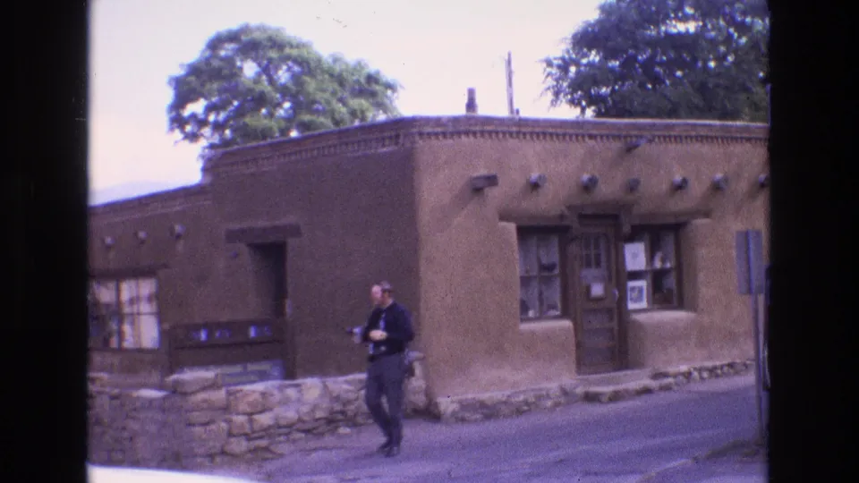 1973 New Mexico home-movie frame showing an adult man in dark clothing walking along a street in front of a brown adobe-style building with wood-framed windows and a stone base.