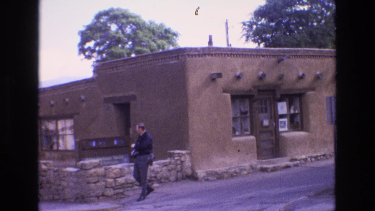 1973 New Mexico frame with an adult man near the curb outside a brown adobe-style exterior, with projecting roof beams, a wooden entrance, and window displays visible.