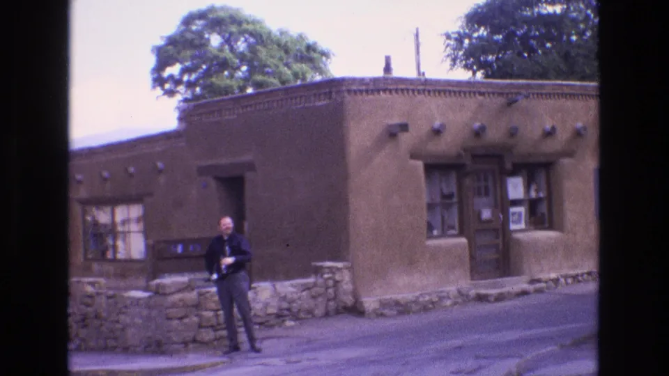 1973 New Mexico frame showing an adult man standing on a street before a brown adobe-style building with a recessed wooden door, display windows, and stone basework.
