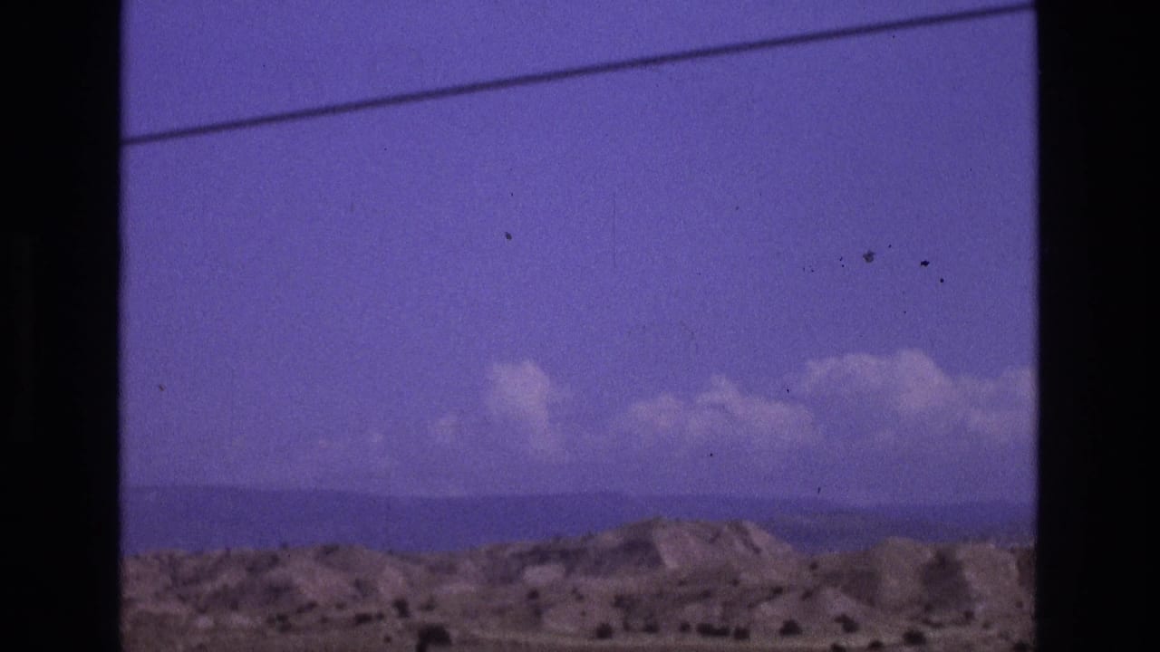 1973 New Mexico 8mm travel frame showing low arid hills, distant horizon, scattered clouds, and a dark vertical border across the view.
