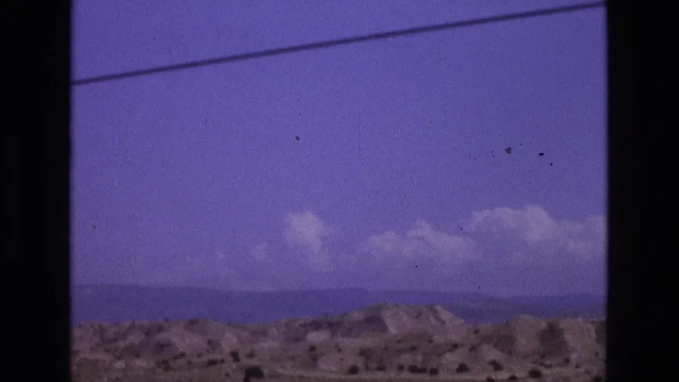 1973 New Mexico 8mm travel frame showing low arid hills, distant horizon, scattered clouds, and a dark vertical border across the view.