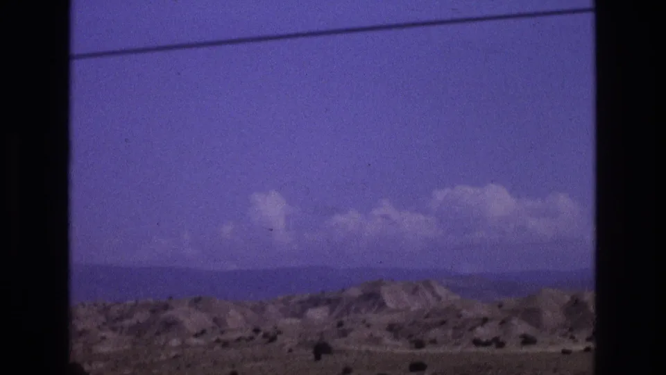 1973 New Mexico 8mm frame with dry rolling hills, a distant horizon, scattered clouds, and a thin overhead power line visible above the landscape.