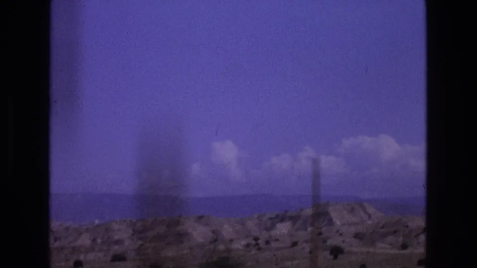 1973 New Mexico travel frame showing a utility pole in front of low desert hills, distant terrain, and a purple-tinted sky with scattered clouds.