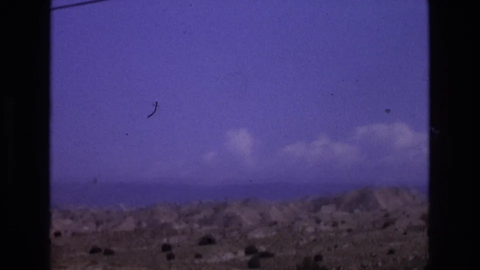 1973 New Mexico 8mm frame showing a broad arid landscape, low hills, scattered brush, distant horizon, and clouds beneath a purple-blue sky.