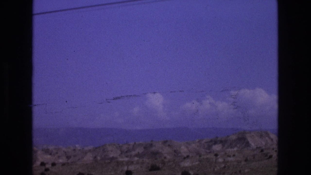 1973 New Mexico desert landscape viewed through a dark window or vehicle frame, with low arid hills beneath a blue-purple sky.