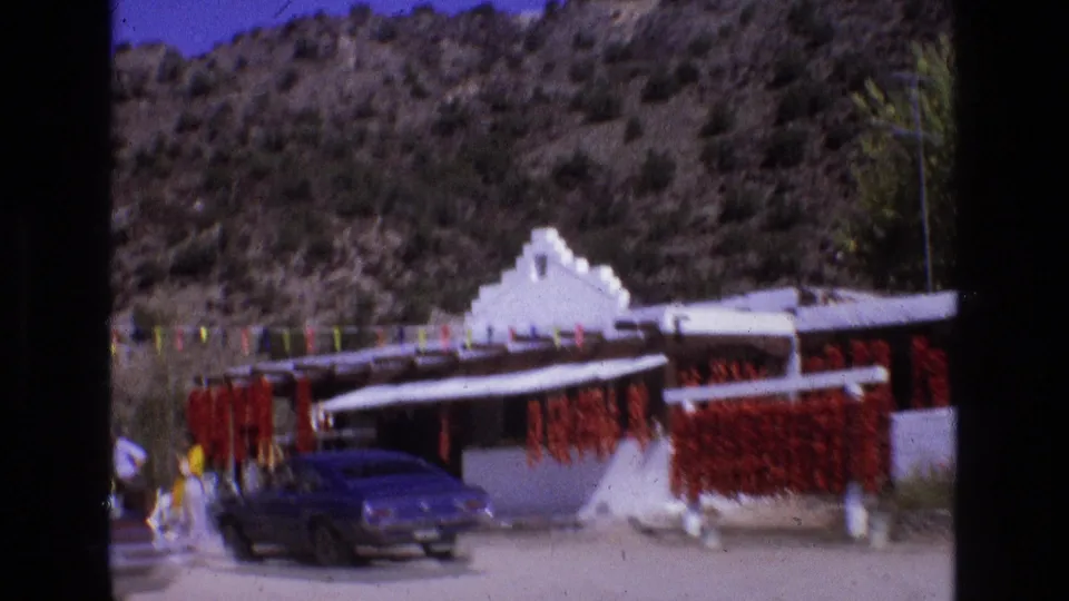 1973 New Mexico roadside market with hanging red chile ristras, colorful pennant flags, a white building, arid hills, and a parked blue classic car.