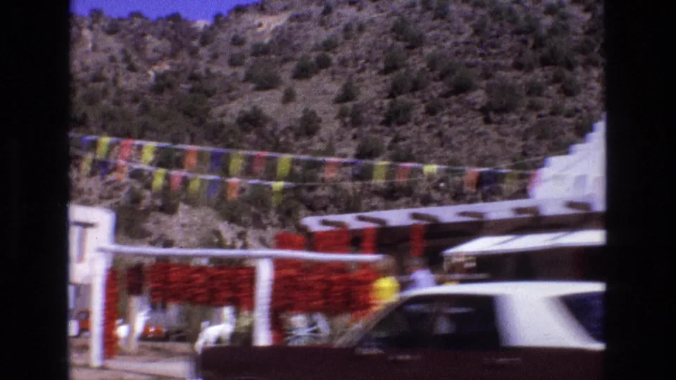New Mexico roadside stand in 1973 with red chile strings, colorful flags, white structures, rocky desert hillside, parked cars, and a small visible person near the market.