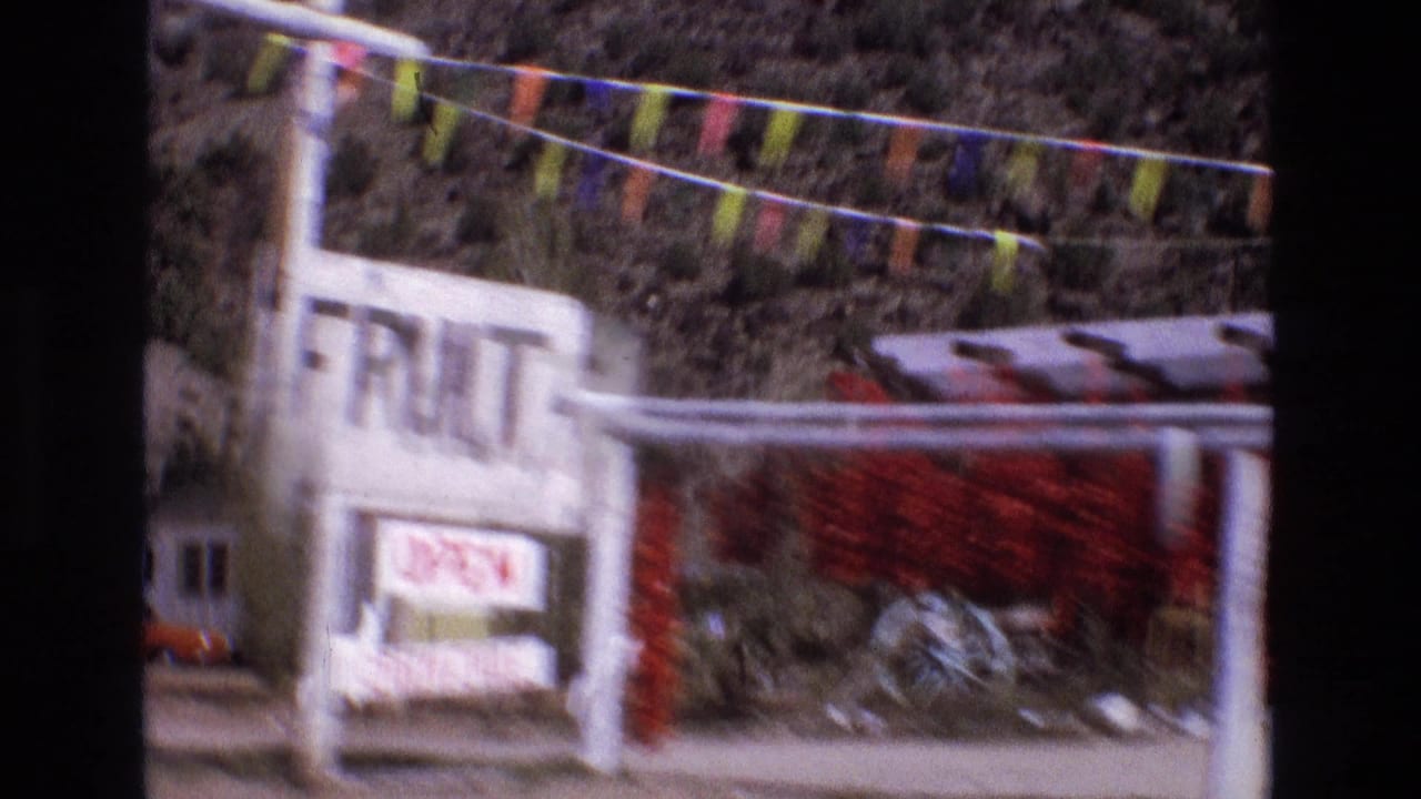1973 New Mexico home-movie frame showing a roadside fruit stand with colorful pennant flags, an OPEN sign, hanging red chiles, and a rocky hillside behind it.