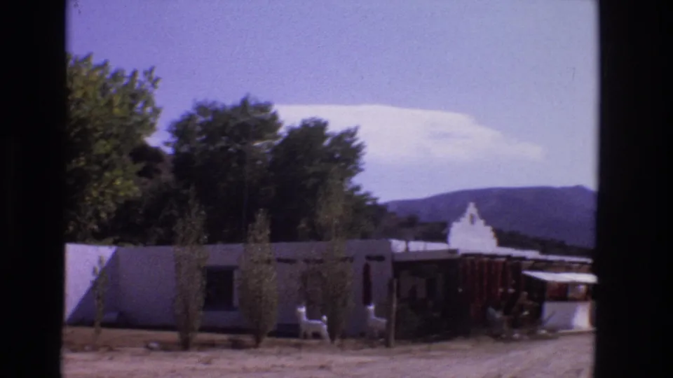 1973 New Mexico home-movie frame showing a white adobe-style building with trees, distant hills, white chairs, and a shaded roadside frontage.