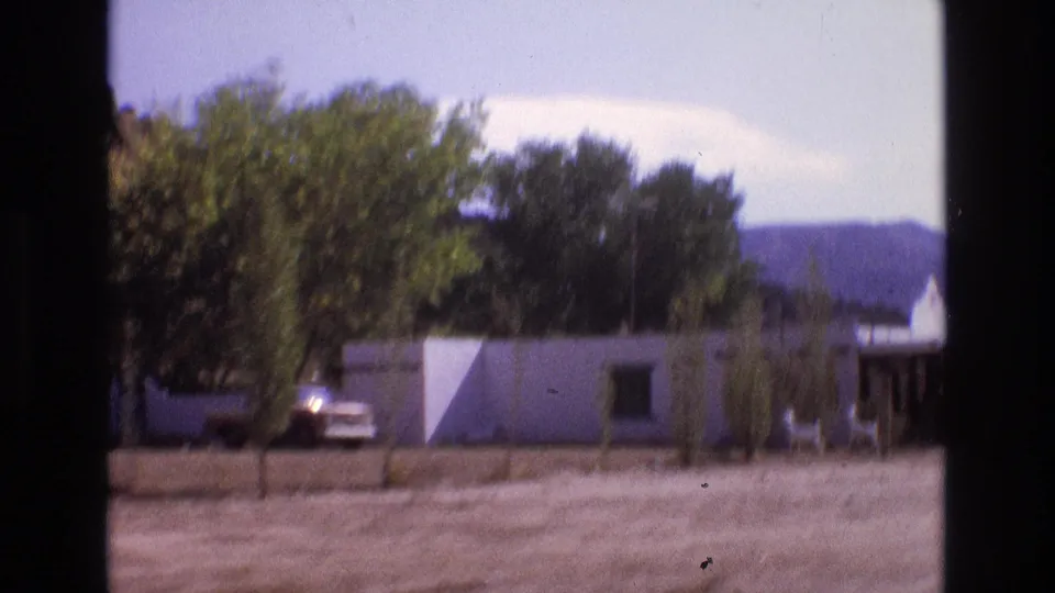 1973 New Mexico frame showing a white roadside building partly shaded by trees, a parked car, dirt frontage, and low hills in the background.