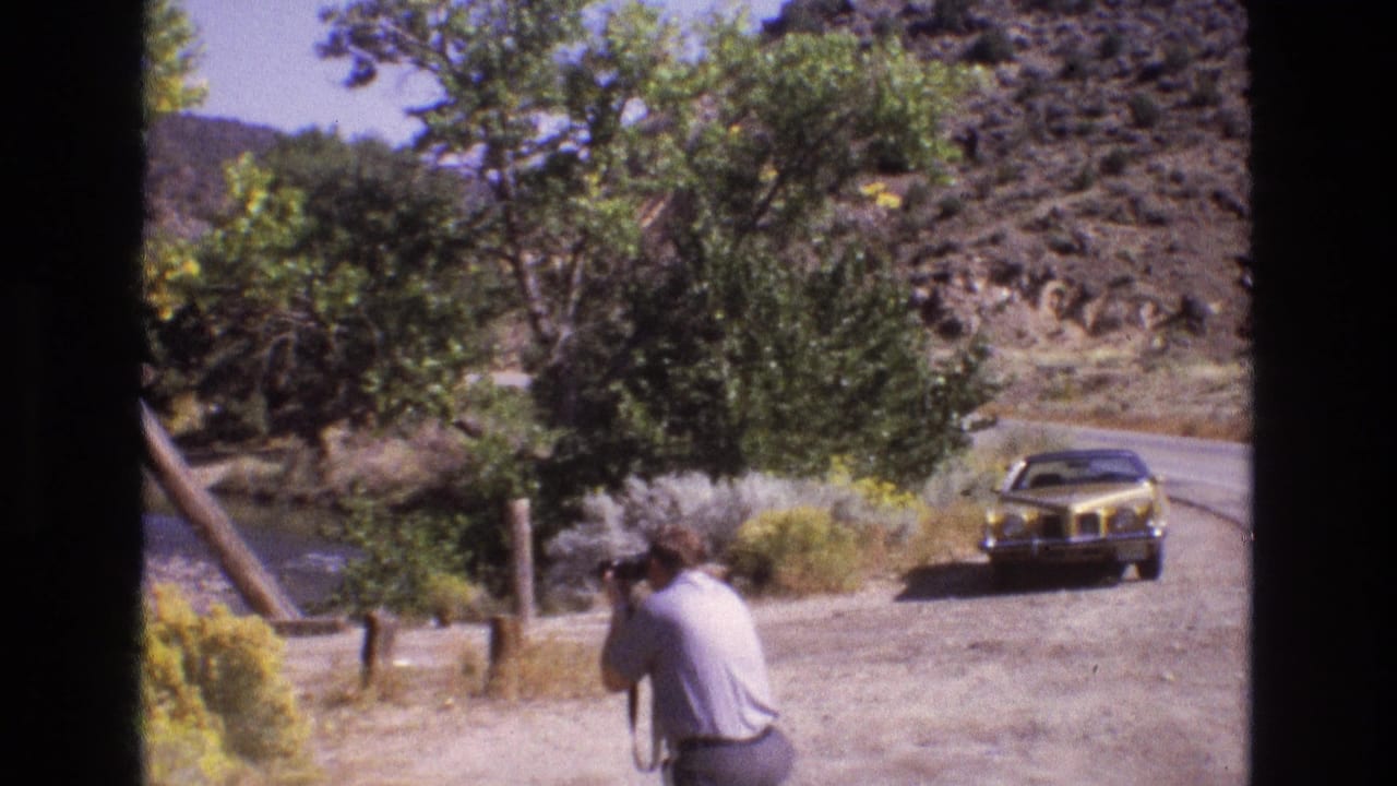 1973 New Mexico 8mm frame showing an adult man with a camera near a dirt roadside, with a yellow car, trees, river edge, and rocky hills in view.