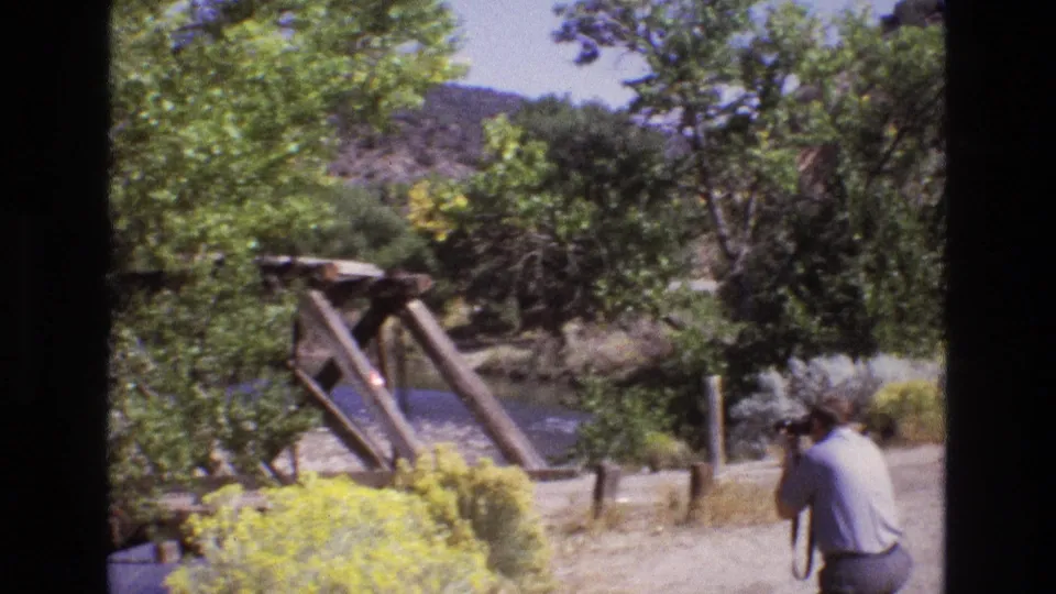 1973 New Mexico 8mm frame with a rustic wooden bridge over a calm river, trees and brush around it, and an adult man partly visible at the roadside.