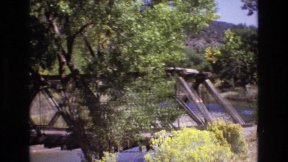 1973 New Mexico 8mm frame showing a weathered wooden truss bridge across a river, surrounded by green trees, yellow roadside brush, and distant rocky slopes.