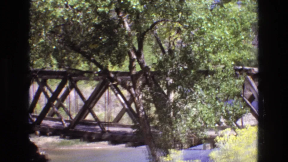 1973 New Mexico 8mm frame centered on the wooden truss bridge crossing the river, partly obscured by trees and bright vegetation in a sunny rural landscape.