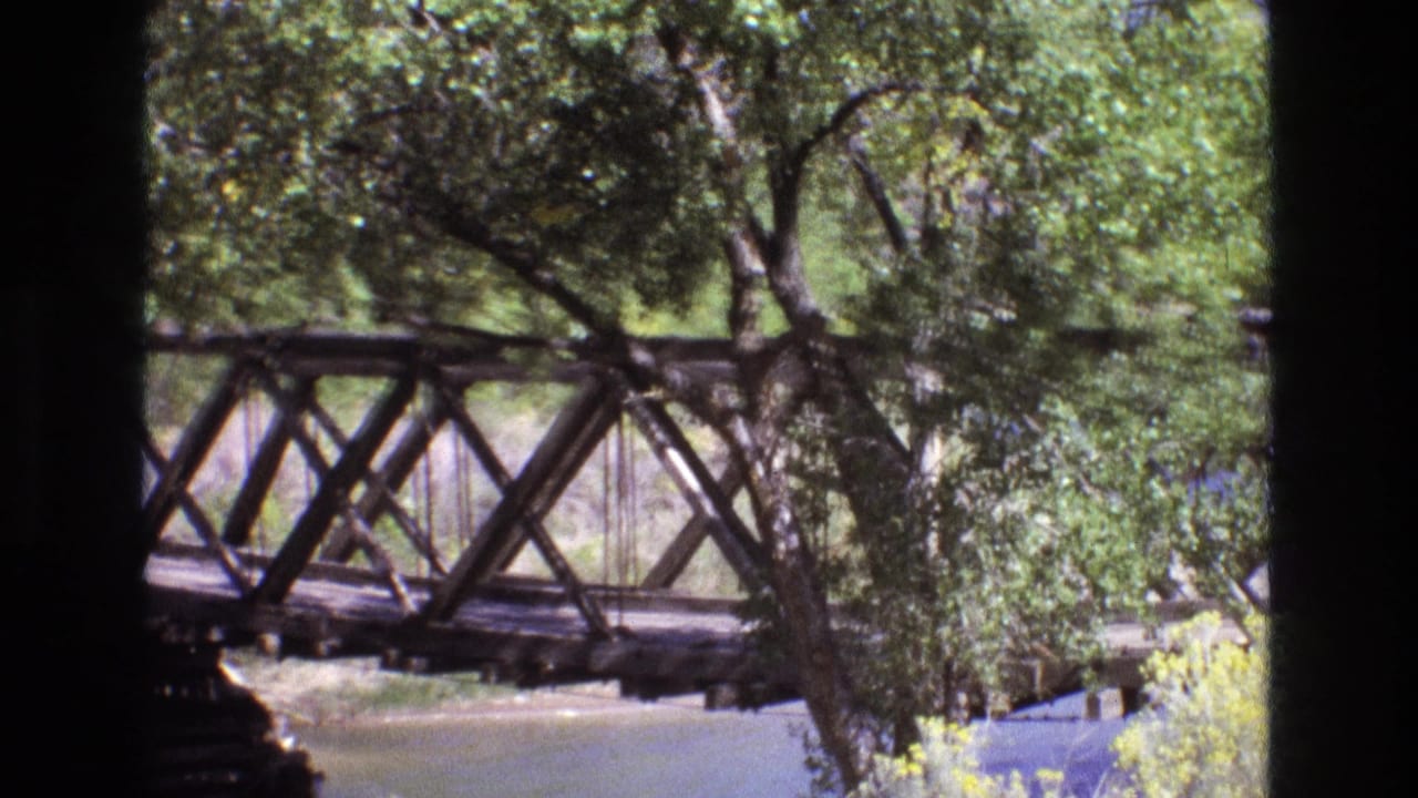 A wooden truss bridge in New Mexico is seen from a distance through leafy trees, with water visible below in 1973 8mm home-movie footage.