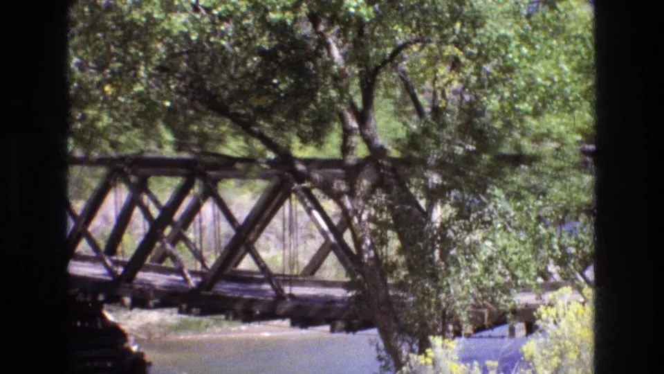 A wooden truss bridge in New Mexico is seen from a distance through leafy trees, with water visible below in 1973 8mm home-movie footage.