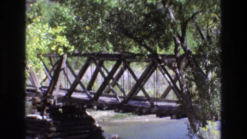 The camera pan brings more of the bridge support and riverbank into view, while trees continue to frame the 1973 New Mexico home-movie scene.