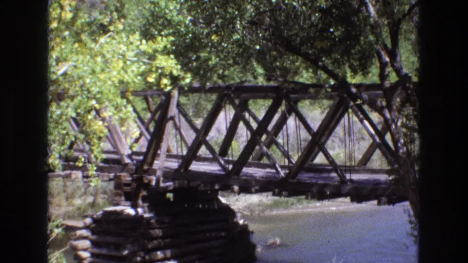 A wooden truss bridge crosses a calm river, with leafy branches and a dark vertical foreground shape partially blocking the New Mexico scene.