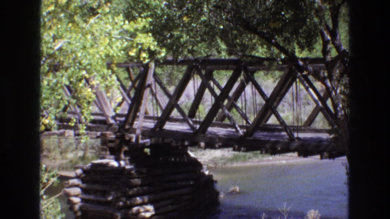 1973 New Mexico 8mm frame showing a weathered wooden truss bridge over a calm river, partly shaded by trees and foliage.