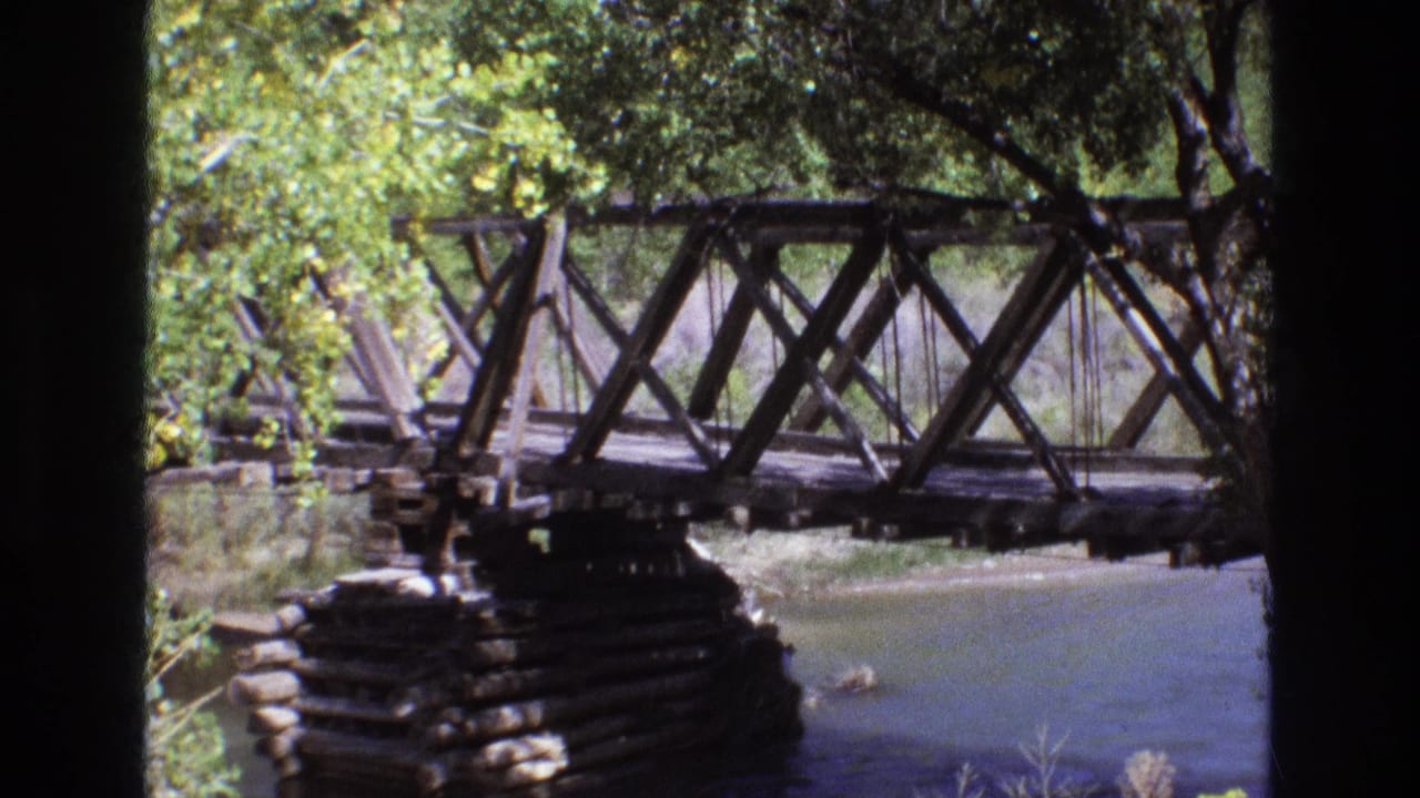 1973 New Mexico 8mm frame with the bridge span centered over still water, framed by leafy trees and dark film-edge shadows.