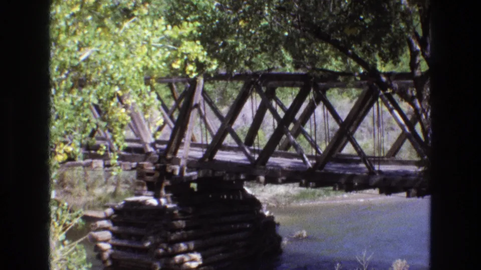 1973 New Mexico 8mm frame showing a closer view of the weathered wooden truss bridge, log support, river surface, and overhanging greenery.