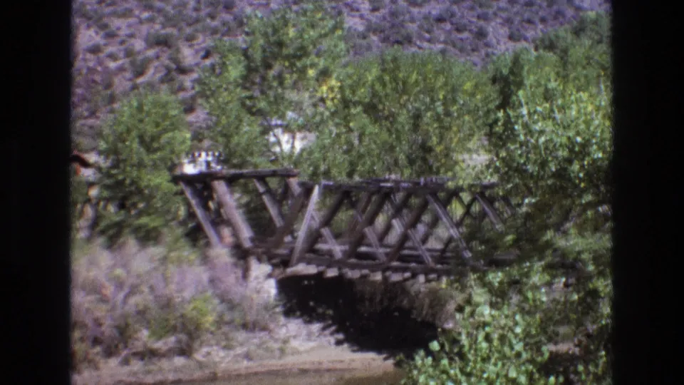 1973 New Mexico 8mm frame showing the bridge from farther back, with trees, brush, dry hillside, and a shaded riverbank around the span.