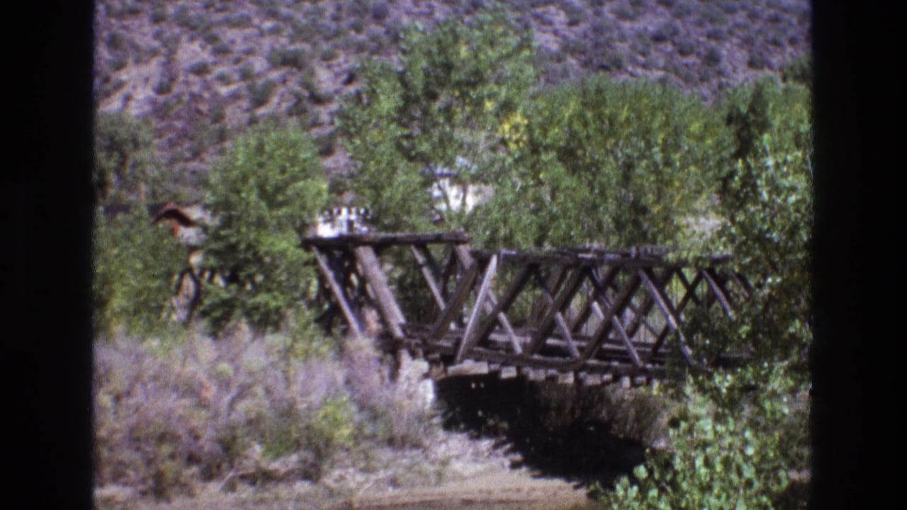 1973 New Mexico 8mm frame showing a rustic wooden truss bridge crossing a dry creek bed with trees and scrub below an arid hillside.